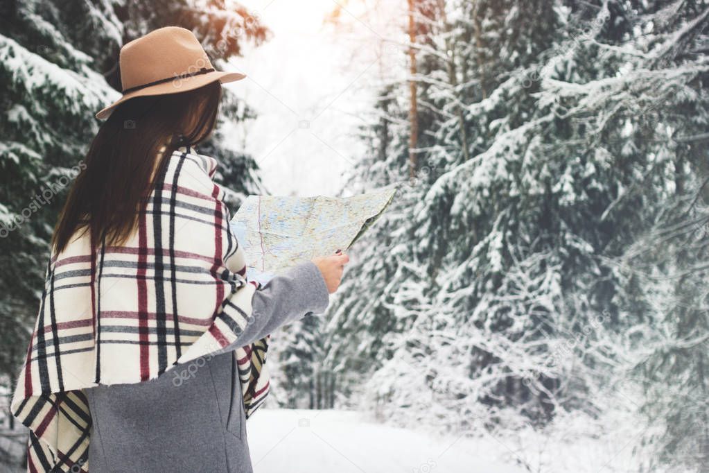 Beautiful woman hold map in hand and traveling around winter forest. Young female hipster walking in snowy landscape. Wearing hat and scarf
