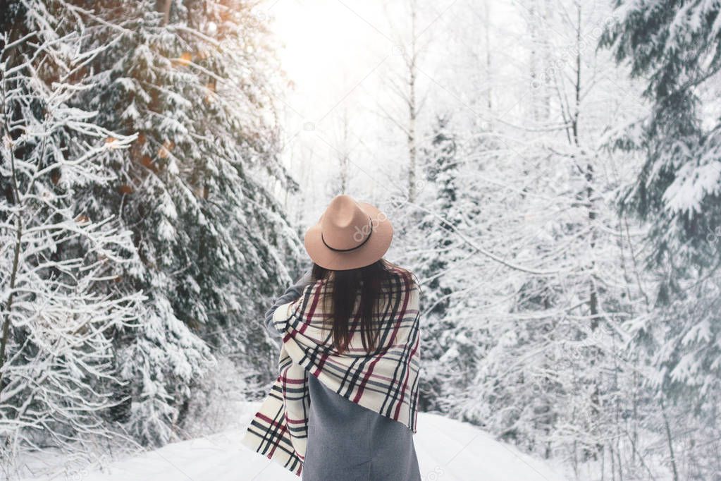 Beautiful woman standing among snowy trees in winter forest and enjoying first snow. Wearing hat, plaid scarf and coat. Wanderlust and boho style