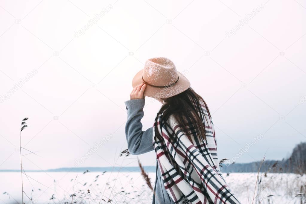 Handsome female hipster holds the hat on her head standing front of amazing natural view on winter lake. Boho style. Wearing vintage hat and plaid scarf