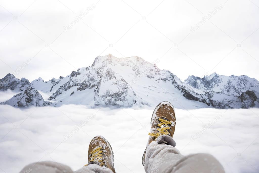 Traveler sitting on mountain peak, feet in hiking boots on background of rocky snowy landscape 