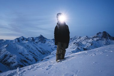 Bir Far konumu muhteşem kış dağlar önünde olan gece explorer görüntüleyin. Cesur Gezgin ile yüksek karlı tepe üzerinde sırt çantası ve snowboard tırmanış. Kayak turu
