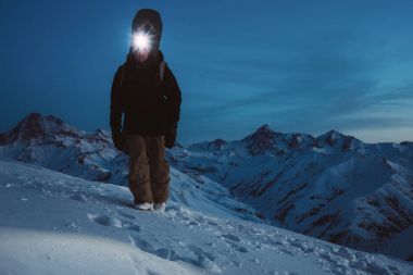 Brave night explorer climb on high mountain. Wearing headlamp, backpack and ski wear. Snowboarder with a snowboard behind his back walking on snowy hill. Backcountry clipart
