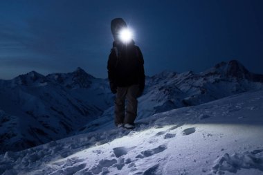 Professional tourist climb on snowy mountain at night and lights the way with a headlamp. Snowboarder walking in front of amazing winter mountains view with backpack and a snowboard behind his back clipart
