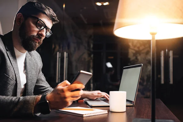 Sakallı işadamı gece loft ofiste cep telefonu kullanarak dizüstü bilgisayar ile. Çağdaş Smartphone'da metin yazarak bir genç adam. Studio coworking, işlem
