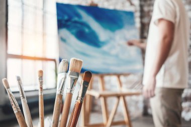 Atölye ile tuval şövale üzerinde çalışan adam ressam. Fırçalar Studio ahşap masa üzerine kapatın. Etkisi flare. Loft alan sanatçı