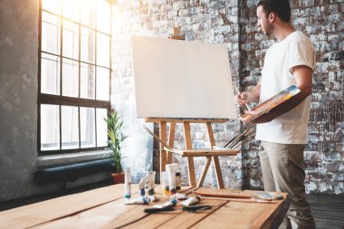 Adam sanatçı çatı sanat stüdyo palette ile şövale üzerinde boş bir tuval önünde duruyor. Erkek ressam boya fırçası elinde tutmak ve boya karıştırın. Hobi kavramı