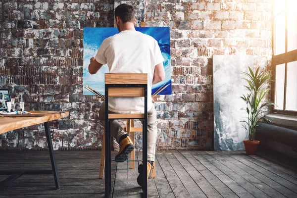 Male painter sits on chair in front canvas and drawing picture in studio. Art class and workshop. Artist painting process. Hobby