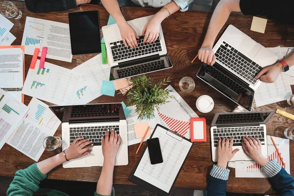 Üstten Görünüm coworking gençlerin dizüstü bilgisayarlar ve kağıt belgeleri üzerinde çalışıyoruz. Grup üniversite öğrencilerinin masa etrafında otururken dizüstü bilgisayar kullanarak. Yeni büyük başlangıç yapma hipsters takım. Kavramı