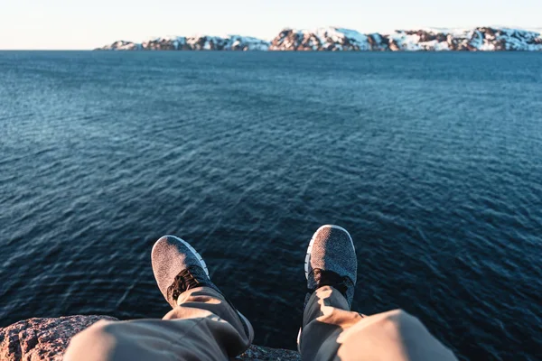 Traveler sitting on cliff in front of breakage above ocean and snow mountains. POV view of legs and shoes on background blue water and rock