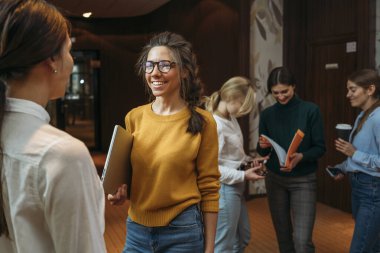 Bir grup mutlu iş kadını modern ofiste toplanıyor. İki pozitif kadın çalışan açık alanda duruyor ve çalışma anını tartışıyorlar.