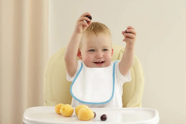 Очаровательный малыш ест абрикос в детском кресле на сером фоне . — стоковое фото