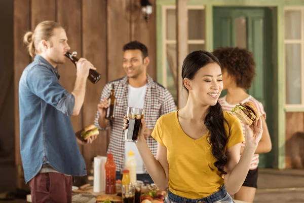 Menina comer hambúrguer com amigos no piquenique — Fotografia de Stock