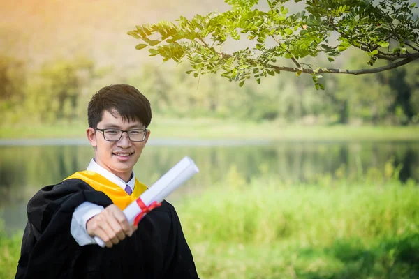 Gelukkig afgestudeerd. Gelukkig Aziatische man in afstuderen toga holdin op nb — Stockfoto