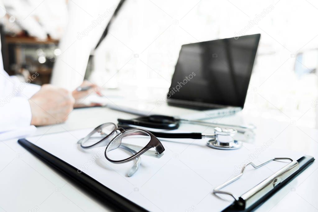 View of stethoscope and equipment on foreground table with doctor using computer keyboard in clinic.