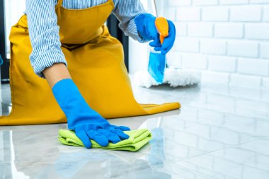 Eş temizliği ve temizlik konsepti mavi eldivenli mutlu genç kadın evde yerleri temizlerken sprey ve toz bezi kullanıyor..