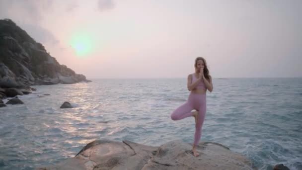 孕妇练习瑜伽 站在海滩上岩石上的Vrksasana的位置上 女孩的手摆出沉思的姿势 女孩的手放在肚子上 向太阳致敬 一个穿着粉色紫色运动服的女人 一个有纹身的女人 冷静点 — 图库视频影像