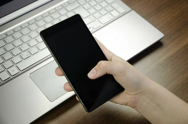 Closeup view of a woman's hand with fingers holding a black smartphone on silver laptop keyboard lying on a wooden mat blurring