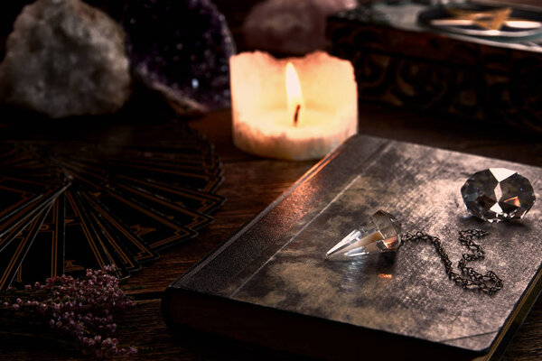 Still life with burning candle on old wooden table top, black fortune teller card, magic book and pentagram box and crystal pendulum. Suitable for magic or esoteric.