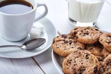 Çikolatalı kurabiyeli tatlı kahvaltı, bir fincan sade kahve ve beyaz tahta masa üzerinde bir bardak süt..
