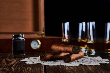 Üç Küba purosu, iki bardak viski ya da rom, çakmak ve higrometreyle dolu ahşap bir kutu. Eski ahşap masa üstü ve siyah arka plan. Metnin için boşluk.