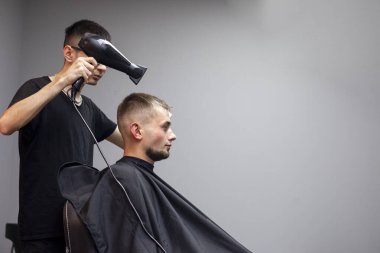 Yakışıklı bir adam berber dükkanında saçını kestiriyor, Kazak bir berber müşterisinin saç kurutma makinesini gri bir duvarda kurutuyor, kuaför kısa saç kesimi yapıyor.