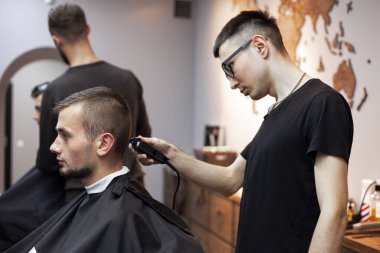 Genç adam berber dükkanında kısa bir saç kesimi yapar, bir berber Kazak bir adamı tıraş makinesiyle keser, aynada yansıtır.