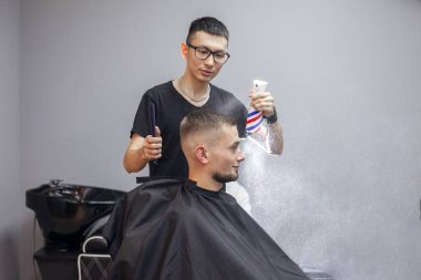 Genç Kazak berber bir berber dükkanında çalışıyor, genç bir adam kuaförde kısa bir saç kesimi yapıyor, kafasını ıslatıyor, arka plandaki gri duvara su sıçrıyor.