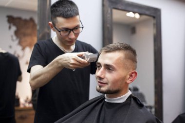 Genç adam berber dükkanında kısa bir saç kesimi yapar, bir berber Kazak bir adamı tıraş makinesiyle keser, aynada yansıtır.