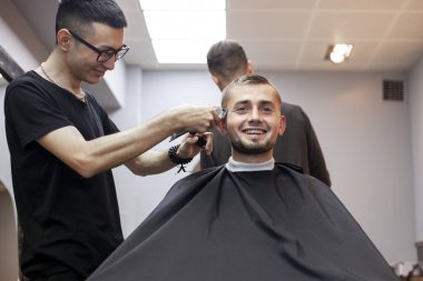 Genç adam berber dükkanında kısa bir saç kesimi yapar, bir berber Kazak bir adamı tıraş makinesiyle keser, aynada yansıtır.