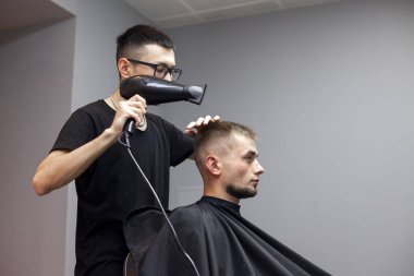 Yakışıklı bir adam berber dükkanında saçını kestiriyor, Kazak bir berber müşterisinin saç kurutma makinesini gri bir duvarda kurutuyor, kuaför kısa saç kesimi yapıyor.