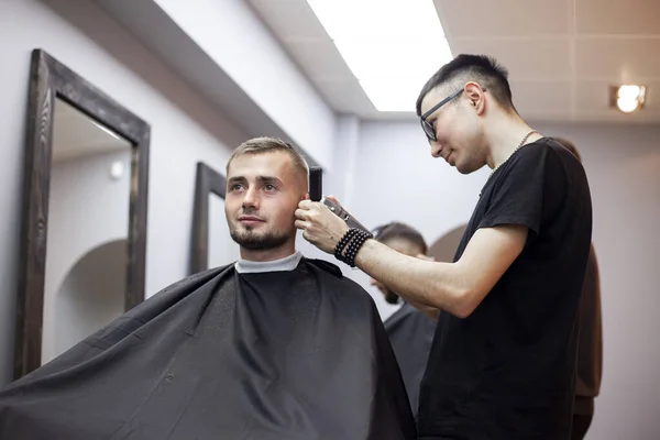young guy makes a short haircut in a barbershop, a barber Kazakh cuts a man with a trimmer, reflection in the mirror