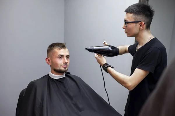 handsome guy has a haircut in a barbershop, a Kazakh barber dries a client\'s hair dryer against a gray wall, a hairdresser makes a short haircut