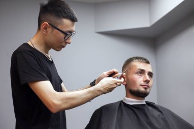 Bir adam berber dükkanında saçını kestirir, genç bir Kazak berber manuel olarak makas ve tarak kullanır, usta kuaförde kısa kestirme yapar.