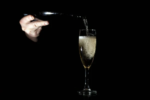 Mão derrama champanhe em um copo em um fundo preto, o barman derrama álcool em uma noite festiva — Fotografia de Stock