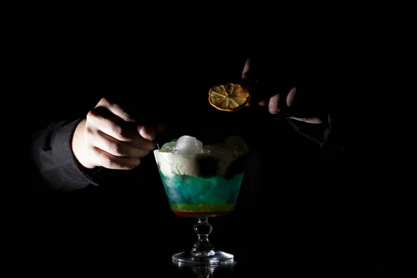Garçom faz um coquetel multi-colorido com gelo e espuma em um copo em um fundo preto, as mãos segurar a bebida — Fotografia de Stock