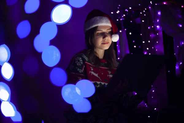 girl student sitting alone for the new year with a laptop at home in Christmas clothes and prints a message, woman online for Christmas