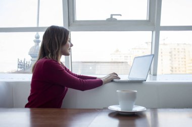 Genç kız bir kafede dizüstü bilgisayarla oturuyor kahve içiyor ve çalışıyor. Sabahları pencerenin yanında bilgisayar kullanıyor.