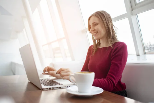 Jovem freelancer menina sentada com um laptop em um café trabalhando e bebendo café, estudante usar um computador de manhã perto da janela — Fotografia de Stock