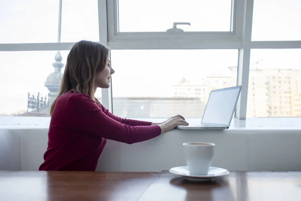 Jovem freelancer menina sentada com um laptop em um café trabalhando e bebendo café, estudante usar um computador de manhã perto da janela — Fotografia de Stock