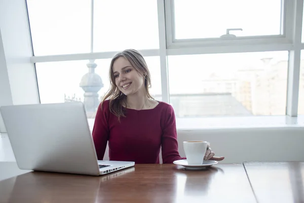 Jovem freelancer menina sentada com um laptop em um café trabalhando e bebendo café, estudante usar um computador de manhã perto da janela — Fotografia de Stock