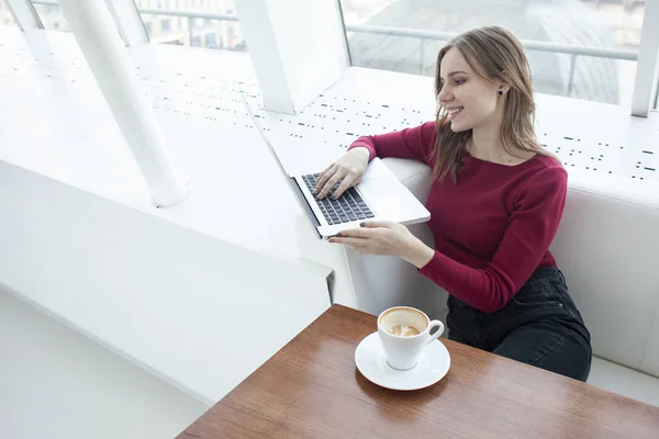 Jovem freelancer menina sentado em um café com um laptop e beber café, mulher usar um computador perto da janela — Fotografia de Stock