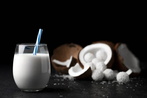 Біле кокосове молоко в склянці на темно-чорному тлі, цукерки з кокосовими пластівцями, продукти для приготування їжі — стокове фото