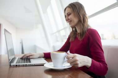 Genç kız bir kafede dizüstü bilgisayarla oturuyor ve kahve içiyor. Kadın pencerenin yanındaki bilgisayarı kullanıyor.
