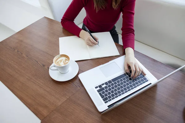 Jovem freelancer menina senta-se em um café com um laptop e escreve em um caderno, um estudante estudando no computador pela manhã — Fotografia de Stock