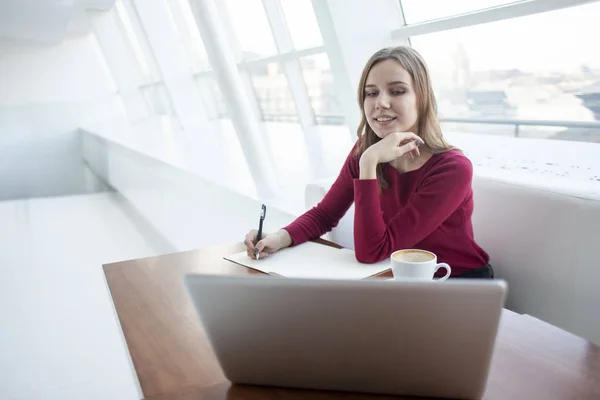 Jovem freelancer menina senta-se em um café com um laptop e escreve em um caderno, um estudante estudando no computador pela manhã — Fotografia de Stock