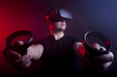 VR gözlüklü bir oyuncu gece oyun odasında bir tetikçiyi oynar, sanal dünyada oyun kumandalarıyla ateş eden bir adam.