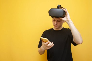 Sarı arka planda VR gözlüklü genç adam akıllı telefon ve gülümseme kullanıyor, oyuncu telefonu sanal gerçekliğe bağlıyor.