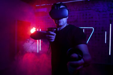 VR gözlüklü bir oyuncu gece oyun odasında bir tetikçiyi oynar, sanal dünyada oyun kumandalarıyla ateş eden bir adam.
