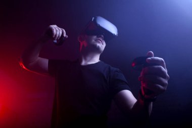 VR gözlüklü bir oyuncu gece oyun odasında bir tetikçiyi oynar, sanal dünyada oyun kumandalarıyla ateş eden bir adam.