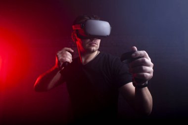 VR gözlüklü bir oyuncu gece oyun odasında bir tetikçiyi oynar, sanal dünyada oyun kumandalarıyla ateş eden bir adam.